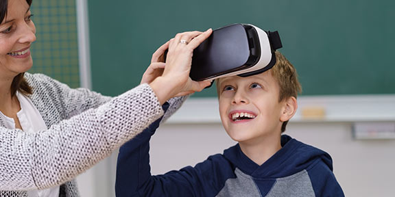 A young boy smiles as his teacher helps him put on a VR headset